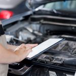 Certified mechanic in garage using tablet to follow checklist while doing maintenance on car. Close up shot of specialist in auto repair shop doing checkup on vehicle engine